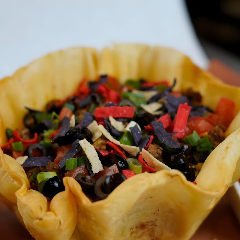 A taco salad in a crispy tortilla bowl, topped with ground beef, olives, tomatoes, and colorful tortilla strips.