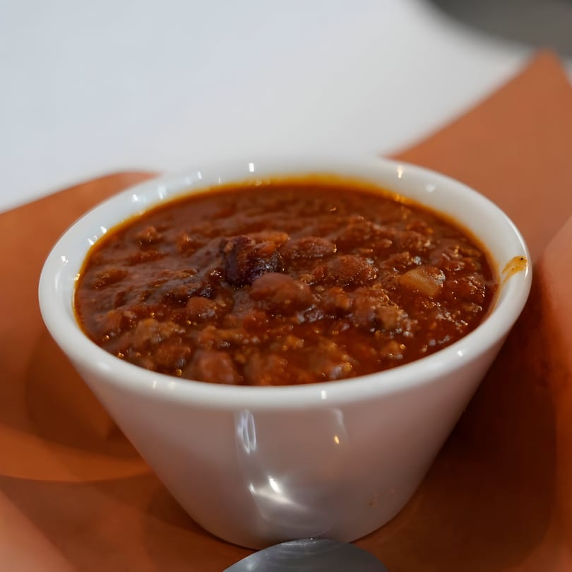 A white bowl filled with hearty chili, featuring beans and ground meat, on a brown surface.